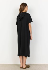 Schwarzes Kapuzenkleid aus weichem Stoff, mit kurzen Ärmeln und Seitenschlitzen, das bis zur Mitte der Waden reicht. Getragen mit braunen Schlupfschuhen.