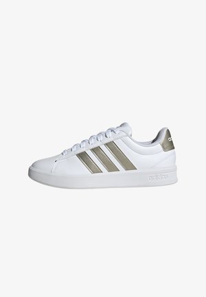 Witte atletische sneaker met drie metalen gouden strepen, een bovenwerk van glad leer, een ronde neus en een vlakke rubberen zool. Branding op de hiel.