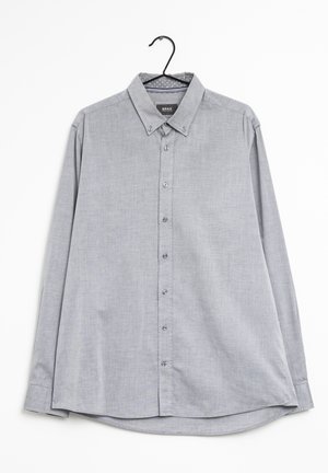 Chemise gris clair à manches longues, boutonnée, avec col boutonné, suspendue sur un cintre noir sur fond blanc.