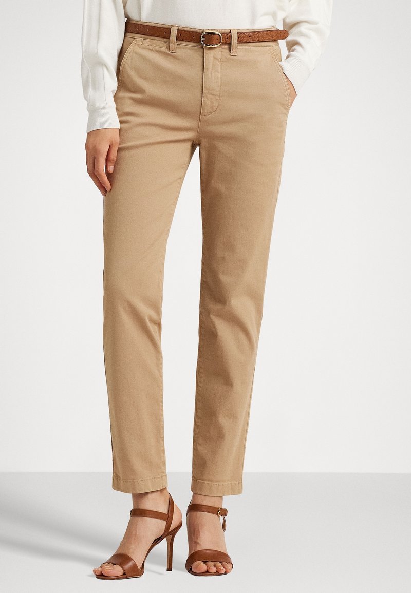 Beige cotton trousers with a straight leg, featuring a brown leather belt and rolled cuffs, paired with brown strappy high-heeled sandals.