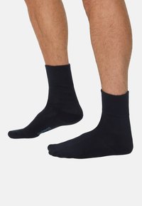 Sorte ankelsokker lavet af en glat stof, med en ribbet kant og en lyseblå logo på undersiden. Sokkerne dækker anklen.