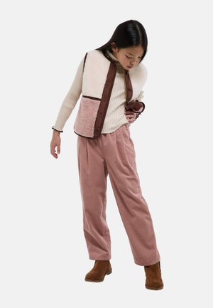 Jeune femme portant un pull crème côtelé, un pantalon large rose, un gilet en peau retournée bicolore et des bottines marron, regardant vers le bas avec une main sur la hanche.