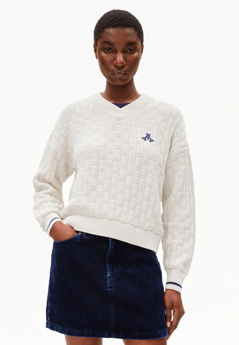 Witte textuur gebreide trui met een V-hals en marineblauwe accenten, gecombineerd met een donkergrode corduroy rok. Voorzien van geribde mouwen en zoom.