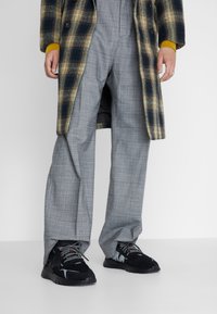 Abrigo de cuadros en marrón y azul, pantalones de cuadros grises, zapatillas negras con cordones estampados. Enfoque en las texturas y colores contrastantes.