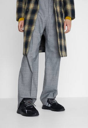 Rutigt överplagg i brunt och blått, grå rutiga byxor, svarta sneakers med mönstrade snören. Fokus på material och kontrasterande färger.