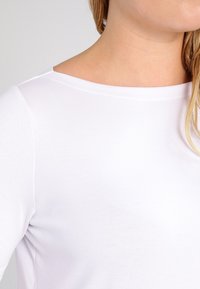 Lähikuva henkilöstä, joka käyttää yksiväristä valkoista pitkähihaista paitaa pyöreällä pääntiellä, näkyvissä on kaula ja osa kasvoista.