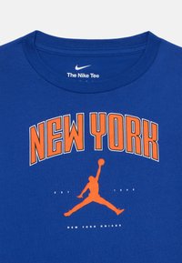 Modré bavlněné tričko s nápisem "NEW YORK" v bílé a oranžové tučné písme, s grafickým siluetovým hráčem v oranžové barvě níže. Kulatý výstřih.