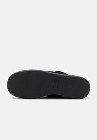 Semelle en caoutchouc noire avec une surface texturée antidérapante, présentant un détail de logo bien visible. Le design est plat et conçu pour une tenue décontractée.