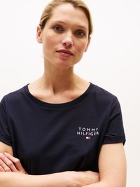 Kvinna med ljus hy och blont hår iklädd en marinblå t-shirt från Tommy Hilfiger, med armarna korsade, som ser lätt åt sidan mot en vit bakgrund.