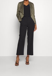 Giacca in suede verde oliva, morbida, con anteriore drappeggiato, indossata sopra un top nero e pantaloni neri a gamba larga; abbinata a tacchi neri con cinturini.