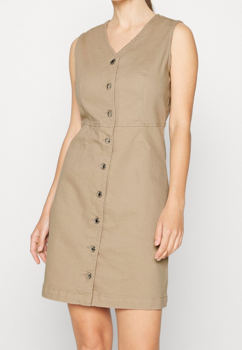 Robe beige sans manches avec fermeture par boutons sur le devant, col en V, taille ajustée et ourlet arrivant aux genoux, portée par une personne debout.