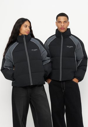 Homme et femme portant des doudounes noires et grises à col montant et pantalons noirs, debout sur un fond blanc uni.