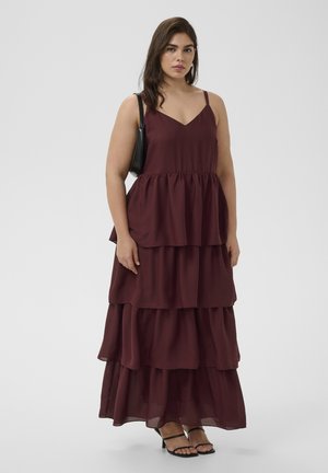 Femme portant une robe maxi bordeaux sans manches à étages avec fines bretelles, des sandales noires à talons et un sac à main noir sur un fond uni.