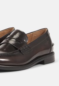 Mocasines de piel en marrón oscuro, con un acabado suave, detalles de costura y un tacón bajo apilado. Diseño clásico con un acento de banda tipo penny.
