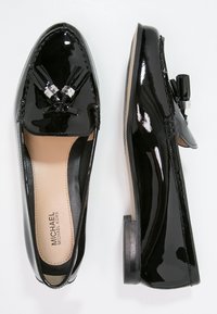 Mocassins en cuir verni noir avec des houppes et des semelles intérieures beiges, présentées en vue de dessus et de côté, avec la marque Michael Kors à l'intérieur.