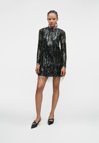 Mujer con el cabello trenzado, vestida con un mini vestido de lentejuelas negro de manga larga y tacones negros de punta, de pie contra un fondo blanco liso.