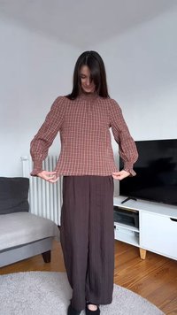 Καφέ μπλούζα με καρό σχέδιο, με φουσκωτά μανίκια και ψηλό γιακά, σε συνδυασμό με φαρδιά, σκούρα καφέ παντελόνια. Υφή υφάσματος με ανάγλυφη όψη και άνετη εφαρμογή.