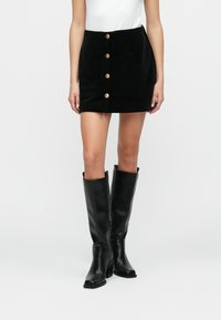 Μαύρη καμισωτή μίνι φούστα με πέντε χρυσά κουμπιά μπροστά, συνδυασμένη με μαύρες δερμάτινες μπότες μέχρι το γόνατο. Απαλή υφή, στενή γραμμή.