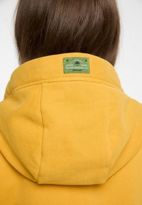 Felpa gialla con cappuccio, dalla finitura testurizzata, con un'etichetta verde sul retro, raffigurante un albero e il nome del marchio.