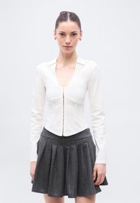 Bijela košulja s dugim rukavima i dubokim V-izrezom, s kopčanjem sprijeda i struktuiranim krojem, uparena sa sivom naboranom suknjom s tankim prugama.
