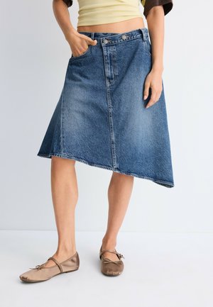 Donna che indossa una gonna asimmetrica di jeans blu, top corto giallo e ballerine beige, in piedi con una mano in tasca.