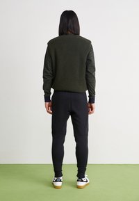 Maslinasto zeleni rebrasti pulover s prugastim rukavima, u kombinaciji s crnim sužavajućim trenirkama i bijelim Nike tenisicama s crnim akcentima.