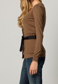 Brauner gestrickter Cardigan mit langen Ärmeln, der eine schwarze Stoffgürtel-Taille hat. Über einer blauen Jeans getragen, die ein körpernahes Design zeigt.