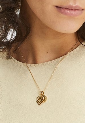 Gold pendant necklace featuring intertwined heart shapes, with a textured chain and smooth surface. Worn over a light-colored top.