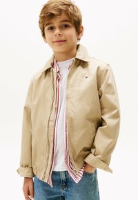 Veste zippée beige avec un col, portée sur une chemise rayée blanche et rouge. Le tissu semble lisse, avec un petit logo sur la poitrine.