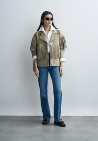 Vrouw met zwarte zonnebril, beige franjesjas, wit overhemd, blauwe spijkerbroek en zwarte loafers, staand tegen een effen muur.