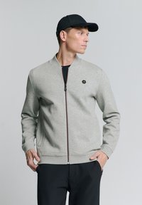 Lichtgrijze zip-up sweatshirt met een staande kraag, ribgebreide boorden en een zwart hexagonaal logo. Draag het over een zwart shirt, met donkere broek.