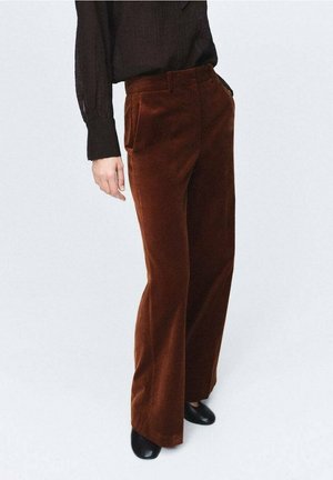 Persona que lleva pantalones marrones de talle alto y pernera ancha, top negro de manga larga y zapatos negros, de pie frente a un fondo claro y liso.