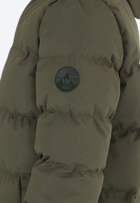 Chaqueta acolchada en verde oliva con un acabado texturizado, que presenta un emblema circular negro en la manga. El diseño está acolchado para una mejor aislación.