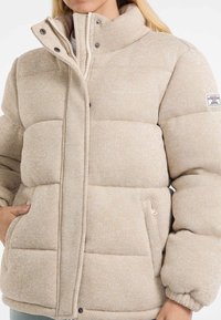 Jachetă puffer bej cu guler înalt, țesătură texturată, nasturi cu cap și buzunare laterale. Are logo-ul "GUESS JEANS" pe piept.