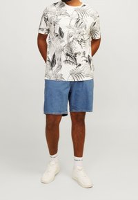 Bílé tričko s krátkým rukávem s černým potiskem tropických listů, doplněné modrými džínovými šortkami. Model má na sobě bílé tenisky a kotníčkové ponožky.