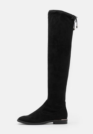Botte plate noire en daim au-dessus du genou, avec un bout arrondi, un petit talon et un lacet ajustable orné de perles métalliques en haut à l'arrière.