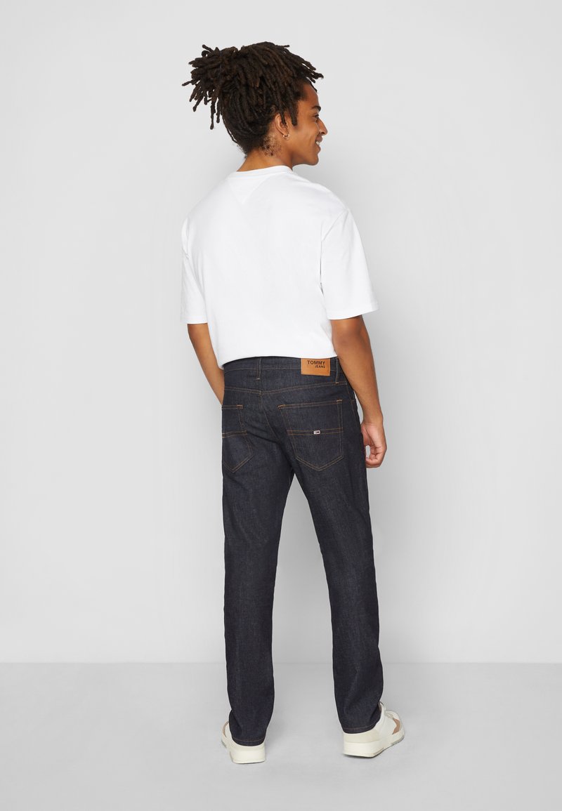 Donkerblauwe denim jeans met een bruine leren label op de achterste tailleband, gecombineerd met een wit T-shirt met korte mouwen en lichte schoenen.
