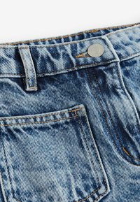 Svetle modré džíny z denimu s vybledlým vzhledem, viditelným švem a kovovým zapínáním. Obsahují přední kapsu a poutko na opasek.