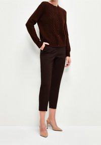 Cardigan en tricot marron avec un motif texturé, associé à un pantalon foncé ajusté et des chaussures à talons nude. Aucuns accessoires visibles.