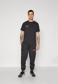 Čierny športový outfit pozostávajúci z trička s nápisom "KING" v zlatej farbe a zodpovedajúcich teplákov s bočnými pruhmi a logotipovými detailmi, vyrobený z hladkej látky.