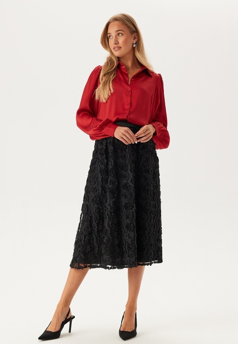 Rode blouse met knopen en lange mouwen; gecombineerd met een zwarte midi-rok van kant met een bloemenpatroon; gedragen met zwarte stilettohakken.