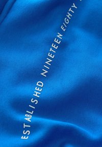 Blauwe stof met de tekst "OPGERICHT NEGENTIEN Tachtig" in het wit, gladde textuur, die de nadruk legt op een levendig, effen kleurontwerp.