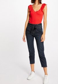 Haut rouge sans manches avec bordure en dentelle, associé à un pantalon en denim foncé taille haute avec ceinture à nouer, et des baskets blanches.