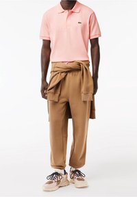 Polo rose clair à manches courtes, pantalons de survêtement marron avec un sweat à la taille, et baskets roses avec des accents foncés.