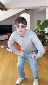 Camisola cinzenta de malha, jeans de ganga azul claro, ténis brancos e óculos de sol transparentes; ambiente interior com plantas e mobiliário.