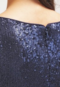 Tmavě modré šaty s flitry s hladkým materiálem s texturovaným povrchem, se zapínáním na zip na zádech a výstřihem ve tvaru lodi.