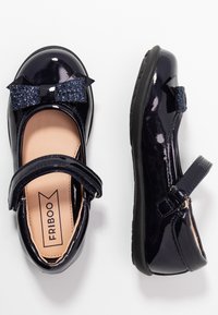 Lakierowane granatowe buty Mary Jane z brokatową granatową kokardą, regulowanym paseczkiem i teksturowaną czarną gumową podeszwą. Zaprojektowane dla wygody i wsparcia.