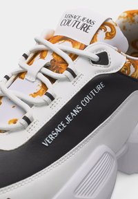 Χοντρό λευκό αθλητικό παπούτσι με μαύρες λεπτομέρειες, περίτεχνο χρυσό μοτίβο κάτω από τα κορδόνια και το λογότυπο "Versace Jeans Couture" στη γλώσσα και στην πλευρική επιφάνεια.
