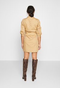 Vestido beige hasta la rodilla con patrones de ojales y mangas abullonadas. Combinado con botas marrones de estampado de serpiente hasta la rodilla. Vista trasera mostrada.