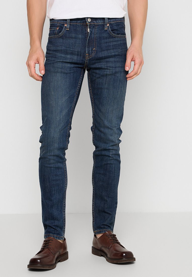 Mężczyzna ubrany w ciemnoniebieskie, slim-fit dżinsy, brązowe skórzane buty eleganckie i białą koszulę, stojący na tle jednolitego białego tła.